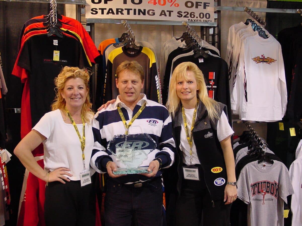 Kelly and Suzy present an award to Doug Sagan of Factory Recreation, standing together at a booth with sportswear and branded shirts.