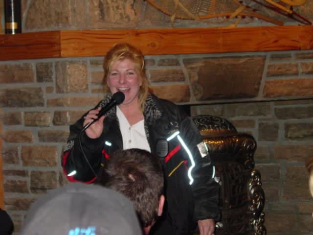 Kelly Shires smiling and speaking into a microphone during a community event, wearing a snowmobile jacket.