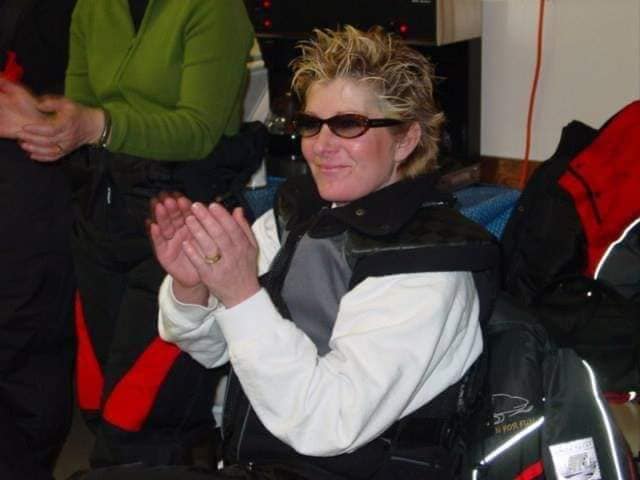 Kelly Shires with sunglasses clapping, dressed in winter hear at an indoor event.